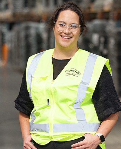 Woman wearing glasses and a Booth's high visibility vest standing, smiling at camera in front of logistics warehouse.